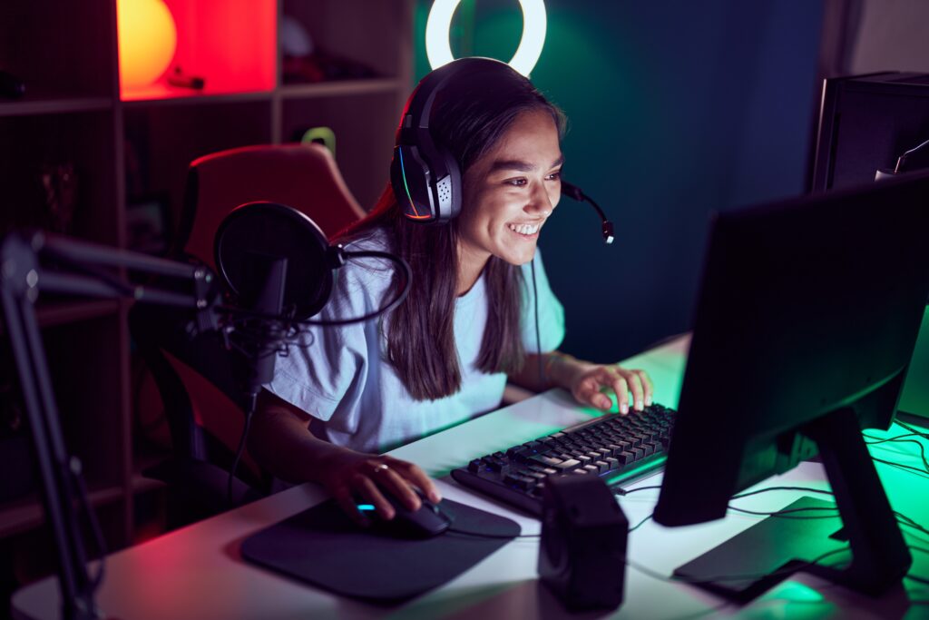 Tenåringsjente spiller dataspill på PC. Hun sitter på en gamingstol og har på seg headset. Hun smiler og ser på skjermen foran seg. Foto:  Krakenimages.com/Adobe Stock