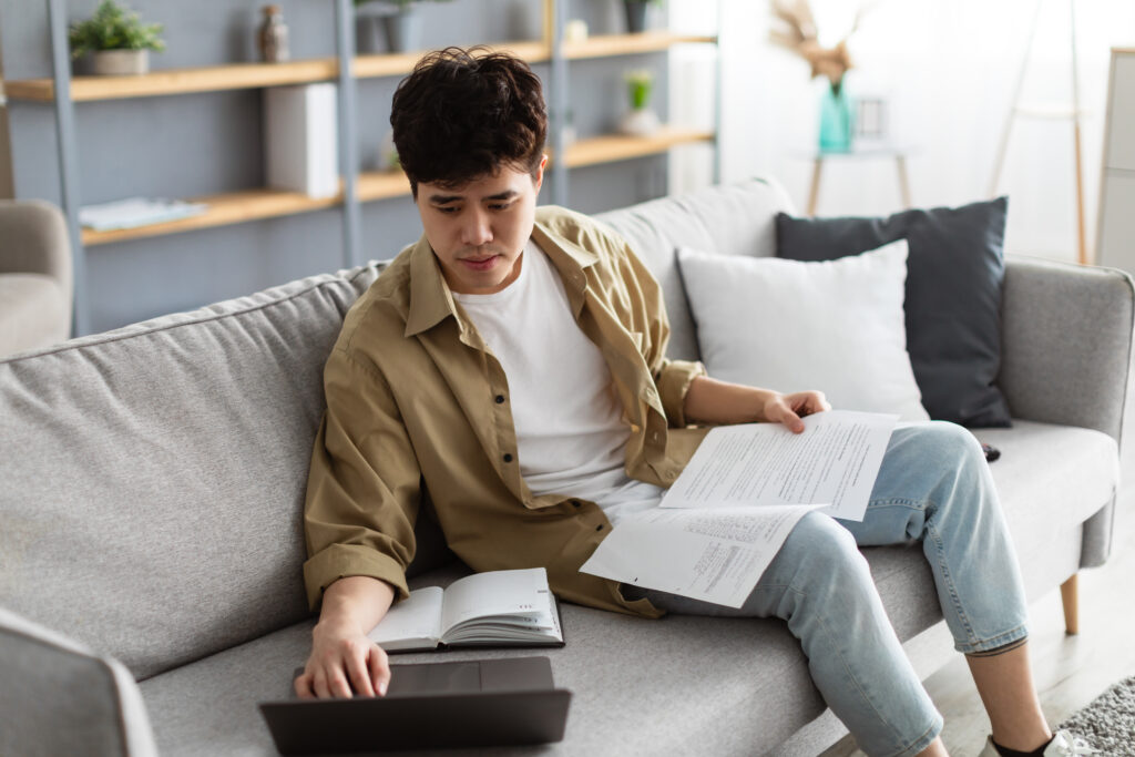 Tenåringsgutt som sitter i sofa med papirer i fanget. Samtidig som han trykker på laptop som står i sofaen. Foto: Prostock-studio/Adobe Stock