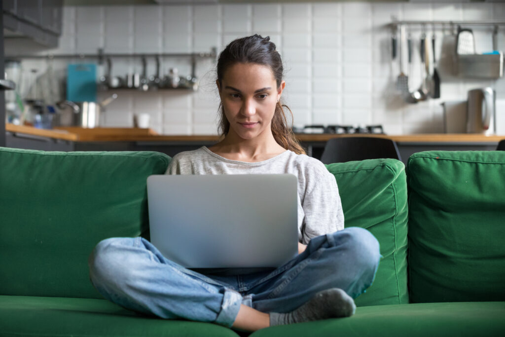 Ung tenåringsjente som sitter i en grønn sofa med en laptop i fanget. Hun kikker til siden og ser betenkelig ut. Bak henne er kjøkkenet.
